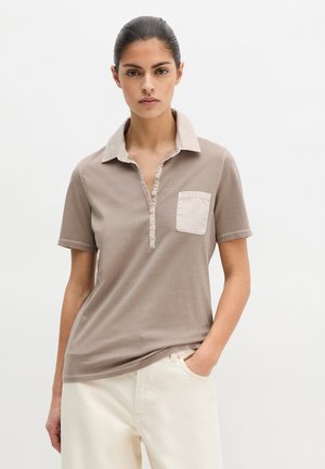 Frau mit zurückgebundenem, dunklem Haar, die ein beiges Poloshirt mit kurzen Ärmeln und Brusttasche sowie weiße Hosen trägt, eine Hand in der Tasche.