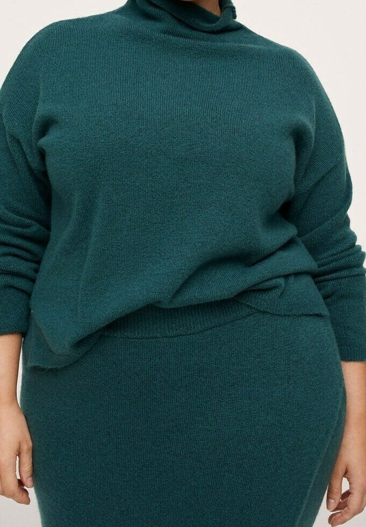 Personne portant un pull en tricot vert sarcelle à manches longues et une jupe assortie en tricot, debout devant un fond uni.