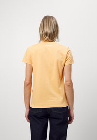 Pomarańczowa bawełniana koszulka z krótkim rękawem, okrągłym dekoltem i małym białym logo z tyłu. Nosi się ją z ciemnymi dżinsami.