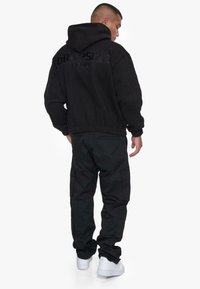 Schwarzer Hoodie mit geprägtem Text "DROPSIZE EST. 2121", kombiniert mit schwarzen Cargo-Hosen und weißen Sneakern. Der Stoff wirkt weich und texturiert.