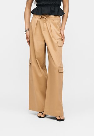 Kvinde iført beige vidde cargobukser med klaplommer og sort ærmeløs bluse med flæser, stående med afslappede arme.