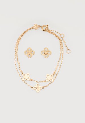 Λεπτή χρυσή αλυσίδα βραχιολιού με τέσσερα μενταγιόν σε σχήμα λουλουδιού και ζευγάρι καρφωτών σκουλαρικιών σε σχήμα λουλουδιού σε λευκό φόντο.