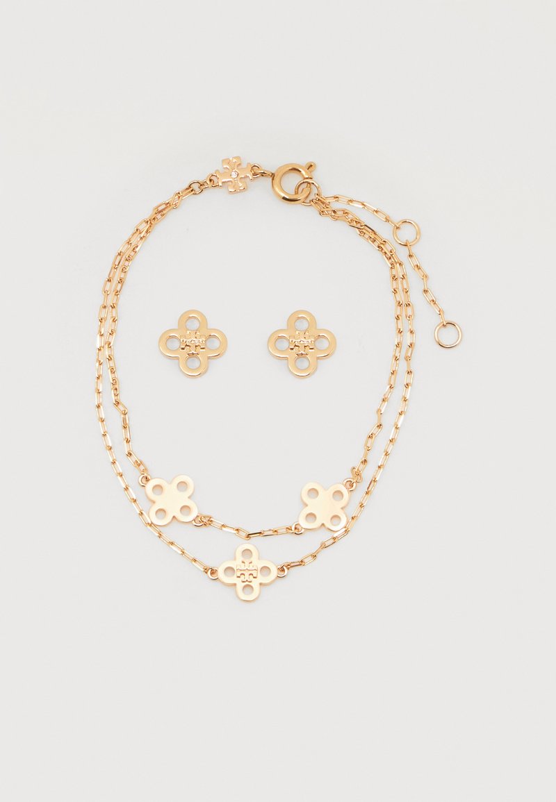 Λεπτή χρυσή αλυσίδα βραχιολιού με τέσσερα μενταγιόν σε σχήμα λουλουδιού και ζευγάρι καρφωτών σκουλαρικιών σε σχήμα λουλουδιού σε λευκό φόντο.