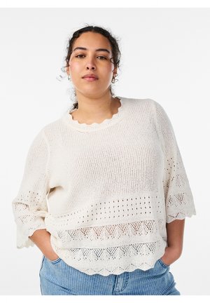 Vrouw met krullend haar die een witte gebreide trui met geschulpte randen en blauwe jeans draagt, staand met handen in de zakken.