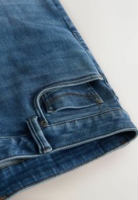 Sinisiä farkkushortsit, joissa on haalistunut pintakäsittely, viisi taskua, kontrastiväriset ompeleet ja kierretty helma. Kupariset niitit korostavat muotoilua.