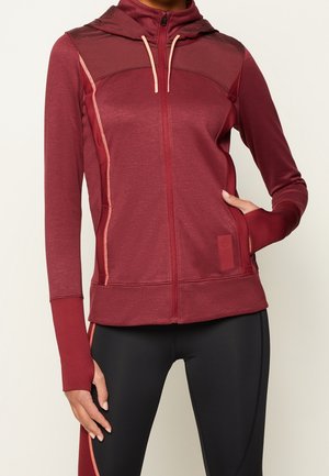 Frau trägt einen kastanienbraunen Kapuzenpullover mit Reißverschluss und Daumenlöchern sowie schwarze Leggings mit kastanienbraunen Streifen, steht mit einer Hand in der Tasche.