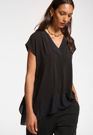 Schwarze V-Ausschnitt-Bluse mit kurzen Ärmeln, die einen geschichteten Saum und eine glatte Textur aufweist. Kombiniert mit schmal geschnittenen schwarzen Hosen. Goldene Statement-Halskette.