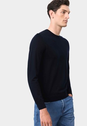 Mand iført en tætsiddende sort langærmet rundhalset sweater og blå jeans, stående med den ene hånd i lommen, neutralt baggrund.