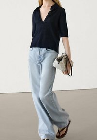 Marineblå strikket kortærmet polo shirt, lyseblå vidde jeans og en lille hvid håndtaske med et tekstureret mønster. Brune flip-flops.