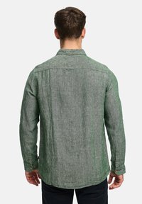 Mand set bagfra iført en langærmet grøn tekstureret skjorte med knapper og mørke bukser mod en ensartet lys baggrund.