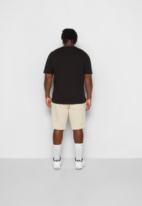 Camiseta negra de manga corta combinada con shorts beige hasta la rodilla. Calcetines blancos y zapatillas deportivas completan el atuendo, visto desde atrás.