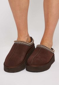 Zoccoli in suede marrone con una finitura testurizzata, caratterizzati da un bordo intrecciato bianco e oro lungo il colletto e una suola spessa e scura.