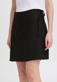 Jupe portefeuille noire en tissu texturé avec une fermeture à nouer à la taille, dotée d'un ourlet droit et de détails de coutures minimaux.