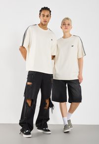 Deux individus portent des t-shirts oversize crème avec des rayures noires sur les côtés. Ils les associent à des jeans noirs déchirés et à des shorts en denim, avec une coupe décontractée.