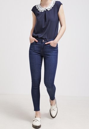 Femme portant un jean slim bleu foncé, un chemisier bleu marine avec un col en dentelle blanche et des chaussures blanches slip-on, marchant les mains dans les poches.