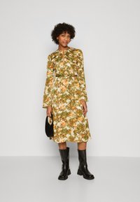 Robe midi florale en tissu vert, orange et blanc avec des manches longues, un décolleté en trou de serrure et une taille nouée, assortie à des bottes noires au genou.