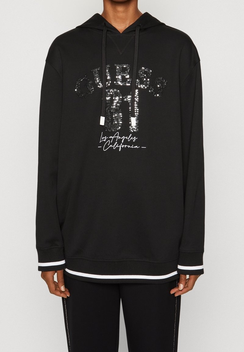 Sudadera negra con un logotipo "GUESS" bordado en lentejuelas y detalles en blanco en los puños y el dobladillo. Presenta una capucha con cordón y un corte holgado.