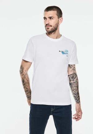 Camiseta de algodón blanca con un estampado gráfico azul y negro en el área del pecho izquierdo, escote redondo y mangas cortas.