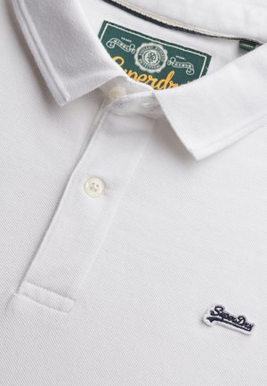 Biała koszulka polo wykonana z teksturowanej tkaniny, z dwudzielnym plisowaniem na dwa guziki oraz granatowym haftowanym logo na lewej piersi. Zawiera zieloną metkę.