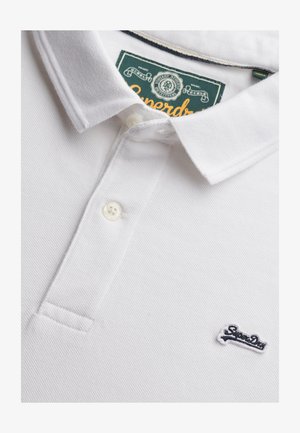 Biała koszulka polo wykonana z teksturowanej tkaniny, z dwudzielnym plisowaniem na dwa guziki oraz granatowym haftowanym logo na lewej piersi. Zawiera zieloną metkę.