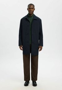 Marineblauer Trenchcoat mit klassischem Design, Knopfleiste und seitlichen Taschen. Über einem grünen Strickpullover und braunen Hosen getragen.