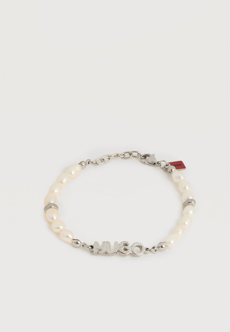 Bracelet avec des perles blanches et des maillons de chaîne en argent, avec des lettres en argent formant "HUGO" et une petite étiquette rouge près du fermoir.