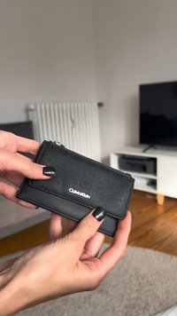 Svart Calvin Klein-lommebok med teksturert overflate, glidelåslomme og en glatt nedre del, holdt i én hånd.