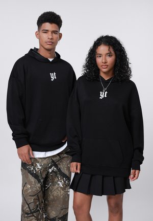 Black hoodies with white "yt" logo, relaxed fit. One model in camo pants, the other in a pleated black skirt. Simple, casual streetwear design.