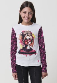 Mädchen mit langen braunen Haaren, das ein weißes Shirt mit rosa blattförmigen Ärmeln trägt, auf dem eine Grafik einer Frau mit Sonnenbrille auf der Vorderseite zu sehen ist.