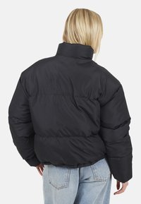 Giacca a vento nera con colletto alto, design trapuntato e vestibilità ampia, abbinata a jeans azzurri chiari. La vista posteriore non mostra hardware visibile.