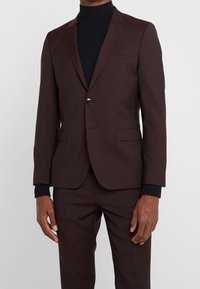 Bordeaux colbert met een enkele knoop, met notch revers en twee voorkeurszakken. Gecombineerd met een zwarte coltrui. Gladde stof, maatwerk pasvorm.