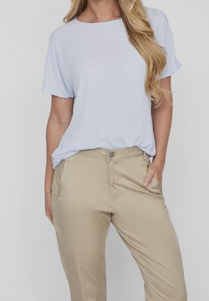 Vrouw draagt een lichtblauw blouse met korte mouwen, ingestopt in beige broek, met handen in de zakken, staand tegen een effen achtergrond.