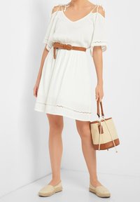 Abito bianco con spalle scoperte, dettagli in pizzo, vita cintrata e cintura intrecciata marrone. Abbinato a espadrillas color sabbia e una borsa a secchiello.