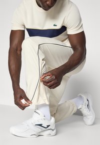 Scarpe da ginnastica bianche con accenti grigi, abbinate a pantaloni sportivi crema con una striscia blu navy e una maglietta a maniche corte abbinata con un logo.