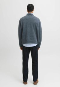 Pull en laine gris avec un col montant, texture tricotée et coupe décontractée, porté sur une chemise blanche. Associé à un pantalon en velours côtelé sombre et des chaussures marron.