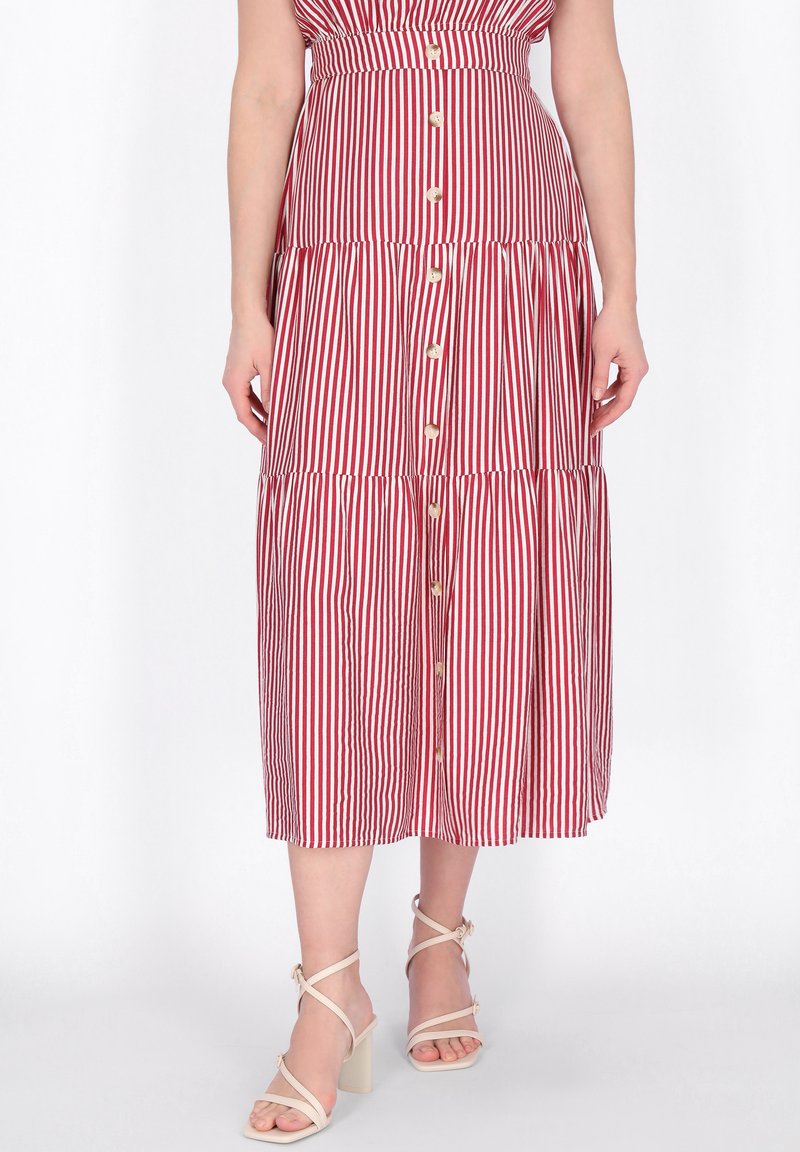 Frau trägt einen rot-weiß gestreiften Midirock mit senkrechten Streifen und Knöpfen vorne, kombiniert mit beigefarbenen, Riemchen-Sandaletten mit hohen Absätzen.