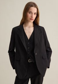 Blazer noir ajusté avec revers, deux poches avant et fermeture à un bouton. Porté par-dessus un gilet noir avec six boutons.