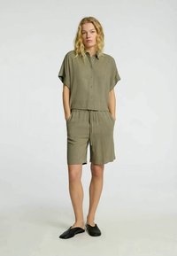 Olivengrøn kortærmet skjorte med knapper parret med matchende shorts. Letvægtsstof, afslappet pasform, sidelommer. Båret med sorte slip-on sko.