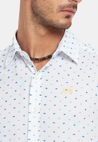 Camisa blanca de botones con puntos azules, un pequeño logotipo amarillo bordado y un cuello en punta. Tejido de algodón con una textura ligera.