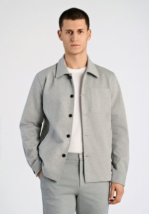 Chaqueta de mezcla de algodón gris con botones negros, cuello estructurado, mangas largas y un bolsillo en el pecho, combinada con pantalones grises a juego.