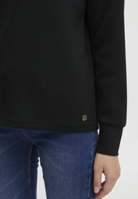 Schwarzes Sweatshirt mit glatter Textur, rundem Ausschnitt und einem kleinen silbernen Logopunkt am unteren Saum. Kombiniert mit blauen Jeans.