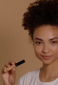 Tube de correcteur KIKO avec un corps beige et un couvercle noir, tenu dans une main. Texture lisse, accents dorés et marquage audacieux sur le côté.