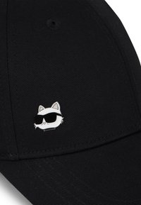 Juodas beisbolo kepuraitė su baltos katės grafika, nešiojančios akinius nuo saulės, kaip segtukas priekyje. Audinys turi tekstūruotą paviršių.