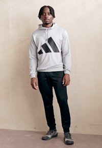 Siva kapucar s puloverom z black Adidas logo, v kombinaciji s črnimi športnimi hlačami in sivimi športnimi čevlji, stoji pred nevtralno podlago.