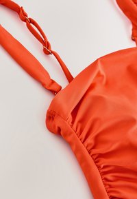 Gros plan d'un vêtement en tissu orange avec des bretelles réglables et des détails de couture froncée sur fond clair.