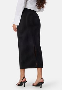 Jupe midi à rayures noires avec fente sur le côté, fabriquée dans un tissu lisse. Portée avec une chemise blanche et des chaussures à talons noires à lanières.