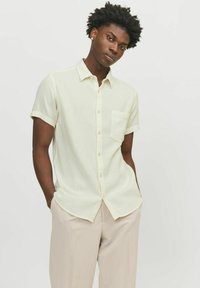 Chemise jaune clair à manches courtes avec un motif texturé, fermeture à boutons à l'avant et une poche poitrine, portée avec un pantalon beige.