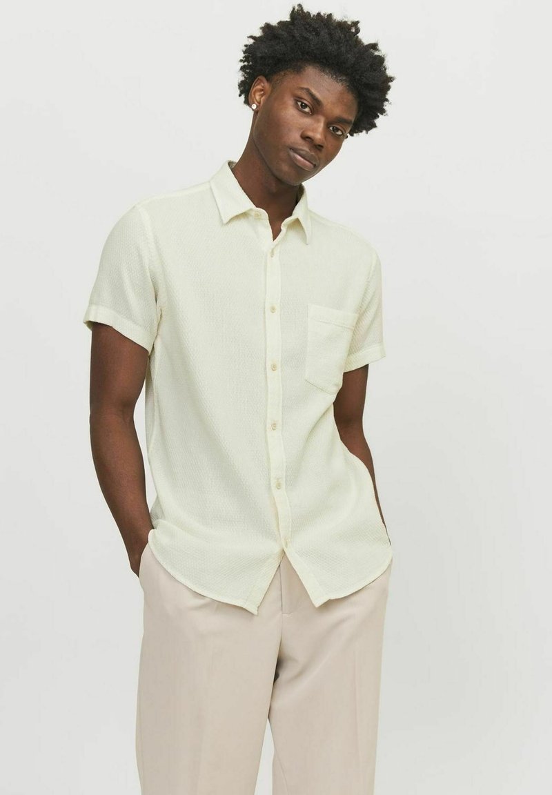 Chemise jaune clair à manches courtes avec un motif texturé, fermeture à boutons à l'avant et une poche poitrine, portée avec un pantalon beige.