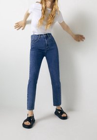 Lekka biała koszulka schowana w wysokich niebieskich dżinsach o prostym kroju i postrzępionym wykończeniu, w połączeniu z czarnymi sandałami na pasku.