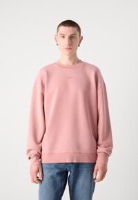 Sudadera rosa de material suave, con cuello redondo, hombros caídos y un sutil detalle del logo en el centro. Combinada con jeans azules.